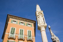 Italy, Veneto, Verona, Pastel painted house facade with pale green window shutters and balconies. Religious statue on free standing pillar in foreground against cloudless blue sky.