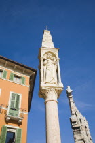 Italy, Veneto, Verona, Pastel painted facade of building with pale green painted window shutters and balconies with religious statue on free standing pillar in foreground against cloudless blue sky.