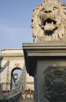 Hungary, Pest County, Budapest, lion statue on Szechenyi Chain Bridge or Memory Bridge with passing pedestrians seen behind.