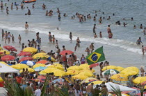 BRAZIL Rio de Janeiro Copacabana beach, crowds on the beach and in the sea, bikinis multi-coloured beach umbrellas and Brazilian flag.