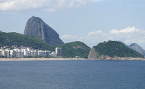 BRAZIL Rio de Janeiro Copacabana beach - hotels and crowds on the beach in the distance, blue sea and sky puffy white clouds, cable car to the top of Sugarloaf Mountain.