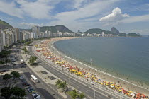 BRAZIL Rio de Janeiro Copacabana, The full sweep of Avenue Atlantica mosiac sidewalk crowds on the beach and in the sea umbrellas hotels tour buses, Sugarloaf Mountain blue sea and sky puffy cloud.