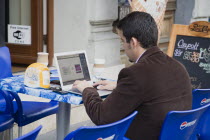 Hungary, Pest County, Budapest, Hungarian man has coffee at outside table while using laptop online with wifi access on Andrassy Ut In Pest. Window sign advertises free internet access and wifi zone.