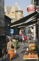 Hungary, Pest County, Budapest, woman walking past local grocer and fruit display under black awning on street lined with parked cars behind Budapest Nyugati palyaudvar rail terminus.