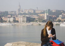Hungary, Pest County, Budapest, young woman using mobile phone on Pest bank of the River Danube with Buda Castle behind.