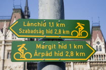Hungary, Pest County, Budapest, green and yellow signs indicating direction of cycle lanes outside the neo-Gothic Parliament Building.