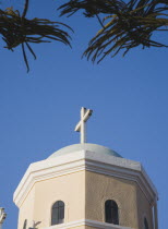 Greece, Dodecanese, Kos. Detail of Greek Orthodox church, hexagonal shape with roof dome and cross, against blue sky, part framed by trees.