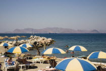 Greece, Dodecanese, Kos, Tigaki Beach. Sunbathers on loungers with blue and yellow striped beach parasols with others swimming in sea and the Turkish coast of Bodrum behind.