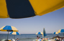 Greece, Dodecanese, Kos. Sunbathers on loungers underneath yellow and blue striped beach parasols on beach outside kos Town.