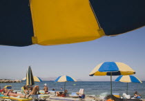 Greece, Dodecanese, Kos. Sunbathers on loungers and blue and yellow striped beach parasols on beach outside Kos Town.
