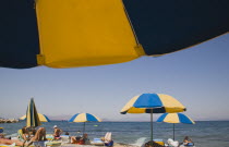 Greece, Dodecanese, Kos. People sunbathing on loungers, some beneath yellow and blue striped parasols on beach outside Kos Town.