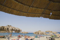 Greece, Dodecanese, Kos, people sunbathing on beach outside Kos Town part framed by thatched shade.