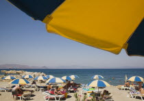 Greece, Dodecanese, Kos. People sunbathing on beach outside Kos Town with sun loungers, blue and yellow striped parasols and people in shallow water of rock enclosed bay beyond. Part framed by undersi...