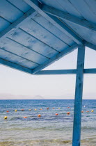 Greece, Dodecanese, Kos. View over red and yellow floats in water part framed by roof of blue painted wooden structure in foreground.