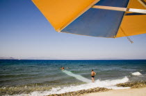 Greece, Dodecanese, Kos, holidaymakers on beach outside Kos Town with blue and orange striped parasol in foreground and man wearing swimming trunks standing at waters edge in cross made by surf lookin...