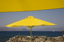 Greece, Dodecanese, Kos, bright yellow parasols on beach outside Kos Town with crumbling stone wall in foreground and view towards distant coastline beyond.