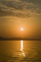 Greece, Northern Aegean, Samos, Vathy, sunset over Samos seen from ferry between Samos and Athens as it leaves Vathy, Samos with gold and orange sky reflected in water and silhouetted landscape.  