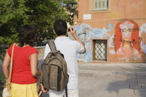 Greece, Attica, Athens, Tourist couple photographing graffiti on building in Plaka district lying just beneath the Acropolis.