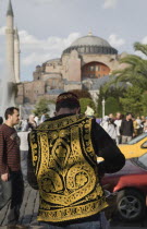 Turkey, Istanbul, Sultanahmet, Man wearing embroidered waistcoat and cap standing at food stall in front of Hagia Sophia part seen beyond.