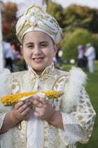 Turkey, Istanbul, Sultanahmet, Young boy eating sweetcorn cooked on the cob, wearing traditional Turkish ceremonial attire standing in front of the Blue Mosque on the day of his circumcision.