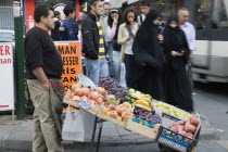 Turkey, Istanbul, Sultanahmet, Two women wearing burqa and veil passing fresh fruit stall with male vendor on busy street beside tramway.