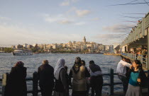 Turkey, Istanbul, Sultanahmet, Crowd leaning on railings beside the Bosphorus admiring view to Galata tower with rods of men fishing from Galata bridge extended out above.