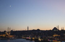 Turkey, Istanbul, Sultanahmet, The Golden Horn, The New Mosque or Yeni Camii at left, the Galata Bridge, and Suleymaniye Mosque at right. Buildings illuminated at dusk and crescent moon in purple, blu...