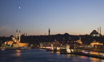 Turkey, Istanbul, Sultanahmet, The Golden Horn. The New Mosque or Yeni Camii at left, the Galata Bridge and Suleymaniye Mosque at right with buildings and restaurants illuminated at dusk and crescent...