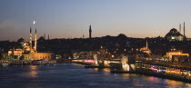 Turkey, Istanbul, Sultanahmet, The Golden Horn. The New Mosque or Yeni Camii at left, the Galata Bridge and Suleymaniye Mosque at right with buildings and restaurants illuminated at dusk and crescent...