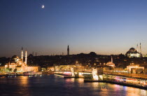 Turkey, Istanbul, Sultanahmet, The Golden Horn. The New Mosque or Yeni Camii at left, the Galata Bridge, and Suleymaniye Mosque at right illuminated at dusk with crescent moon in sky above and coloure...