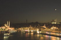 Turkey, Istanbul, Sultanahmet, The Golden Horn. The New Mosque or Yeni Camii at left, the Galata Bridge, and Suleymaniye Mosque at right illuminated at night with crescent moon in sky above.