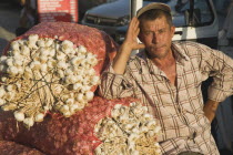 Turkey, Aydin Province, Kusadasi, Stallholder at weekly market leaning on sacks of garlic in the late afternoon summer sunshine.