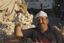 Turkey, Aydin Province, Kusadasi, Stallholder at weekly market holding up bunch of garlic bulbs with more garlic piled up and in sacks behind. Late afternoon summer sunshine.