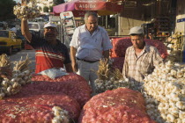 Turkey, Aydin Province, Kusadasi, Stallholder selling garlic  at weekly market standing behind stall stacked high with sacks of garlic bulbs and holding up bunch of tied bulbs in late afternoon sunshi...