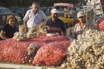 Turkey, Aydin Province, Kusadasi, Stallholder selling garlic at weekly market standing behind stall piled with sacks of garlic bulbs in late afternoon summer sunshine.