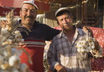 Turkey, Aydin Province, Kusadasi, Stallholders at weekly market with sacks of garlic for sale and holding up bunches of bulbs in late afternoon summer sunshine.