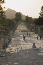 Turkey, Izmir Province, Selcuk, Ephesus, Tourists on paved road lined by broken columns in late afternoon sun in antique city of Ephesus on the Aegean sea coast.