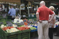 Austria, Vienna, The Naschmarkt, Tourists looking at display on fresh produce stall in front of glass fronted shop front including aubergines, tomatoes, sweetcorn and cauliflower.