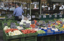 Austria, Vienna, The Naschmarkt, Stallholder standing behind, and with back to, display of fresh produce including tomatoes, sweetcorn, aubergine and mushrooms.