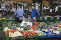 Austria, Vienna, The Naschmarkt, Stallholder and customer standing behind display of fresh produce including sweetcorn, tomatoes, aubergine and mushrooms on stall infront of glass fronted shop.