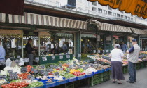 Austria, Vienna, The Naschmarkt, Customers at fresh produce stall with display including peppers, leeks, tomatoes and mushrooms in foreground of glass shopfronts.