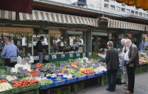 Austria, Vienna, The Naschmarkt, Customers at fresh produce stall looking at display that includes tomatoes, aubergines, leeks, courgettes and mushrooms.