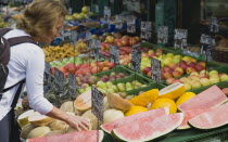 Austria, Vienna, The Naschmarkt, Female customer selecting melon from display on market fruit stall that also includes apples and pineapple.