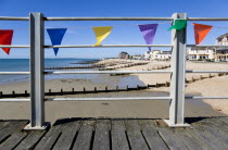England, West Sussex; Bognor Regis, Wooden groynes at low tide used as sea defences against erosion of the shingle pebble beach seen through metal railings with colourful flags on the pier.