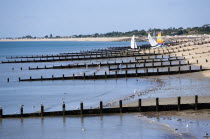 England, West Sussex, Bognor Regis, Wooden groynes at low tide used as sea defences against erosion of the shingle pebble beach with people on the sand by the waterline and small sailing boats on the...