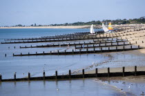 England, West Sussex, Bognor Regis, Wooden groynes at low tide used as sea defences against erosion of the shingle pebble beach with people on the sand by the waterline and small sailing boats on the...