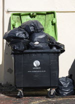 England, West Sussex, Bognor Regis, Wheeled waste bin container overflowing with full large black refuse sacks and bags.