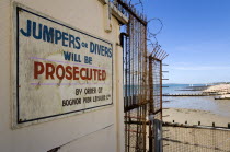 England, West Sussex, Bognor Regis, Warning sign against jumping or diving off the pier into the water on the pier with wooden groynes at low tide used as sea defences against erosion of the shingle p...