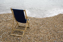 England, West Sussex, Bognor Regis, Single dark blue deck chair on pebble shingle beach looking out to sea with waves gently breaking on the shoreline.