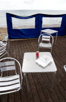 England, West Sussex, Bognor Regis, Empty aluminium chairs and tables on wooden decking behind a wind break on the deserted beach with soft drinks and empty polystyrene food container on table.